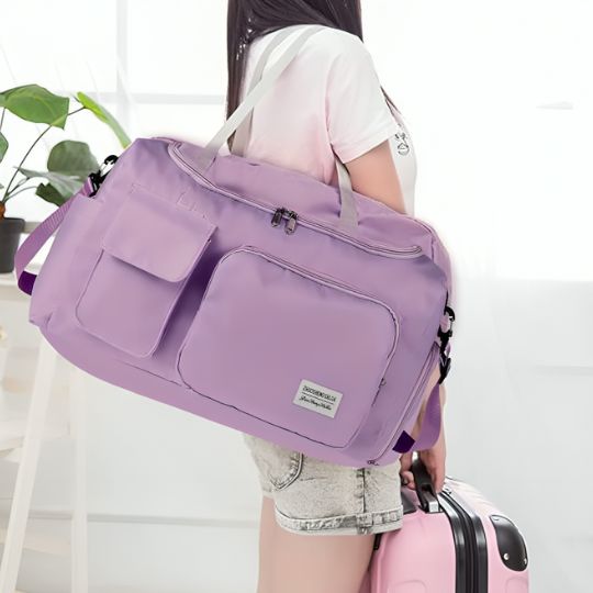Sac de voyage violet porté à l’épaule par une jeune femme, avec une valise à roulettes à proximité.