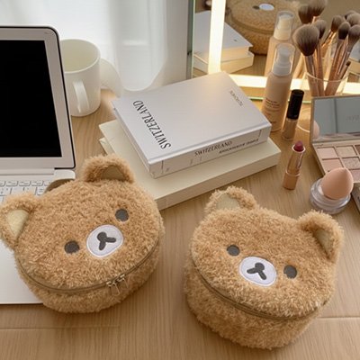 Deux trousses à maquillage beiges en forme d’ours sont posées sur un bureau en bois, à côté d’un ordinateur portable, de livres