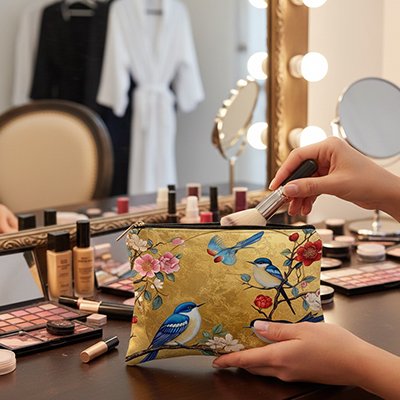 Femme déposant un pinceau dans une trousse maquillage dorée à motifs oiseaux et fleurs, geste délicat sur coiffeuse élégante avec produits beauté
