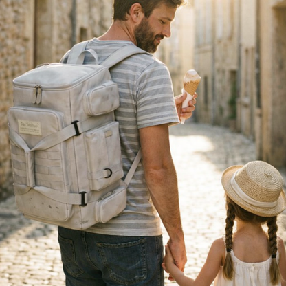 Homme portant un sac à dos de voyage à grande ouverture, blanc, marchant avec un enfant dans une rue pavée ensoleillée