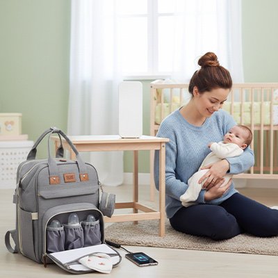 Jeune mère avec bébé dans les bras son mobile est sur la charge avec la prise USB du sac de voyage pour bébé prête pour un départ à l'aventure