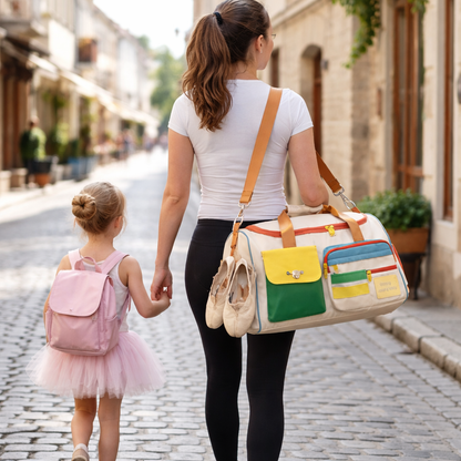 Mère et enfant marchent dans une rue pavée avec un sac crème coloré, chaussures de ballet suspendues, tenue sportive, direction un cours de danse
