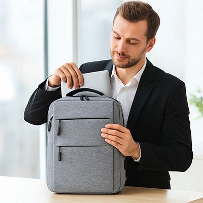 Homme professionnel insérant un ordinateur portable dans un sac à dos gris, posé sur un bureau lumineux, illustrant un usage travail et déplacements.