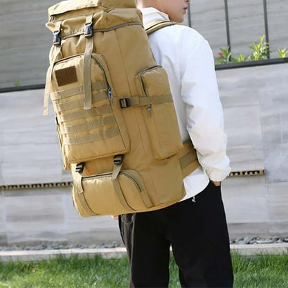 Homme de profil portant un grand sac à dos de voyage beige, épais et structuré, avec nombreuses poches zippées et sangles, idéal pour les déplacements et l’aventure.
