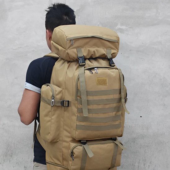 Homme vu de dos portant un grand sac à dos de voyage beige, robuste et volumineux, avec multiples poches et sangles, adapté à la randonnée et aux longs déplacements.