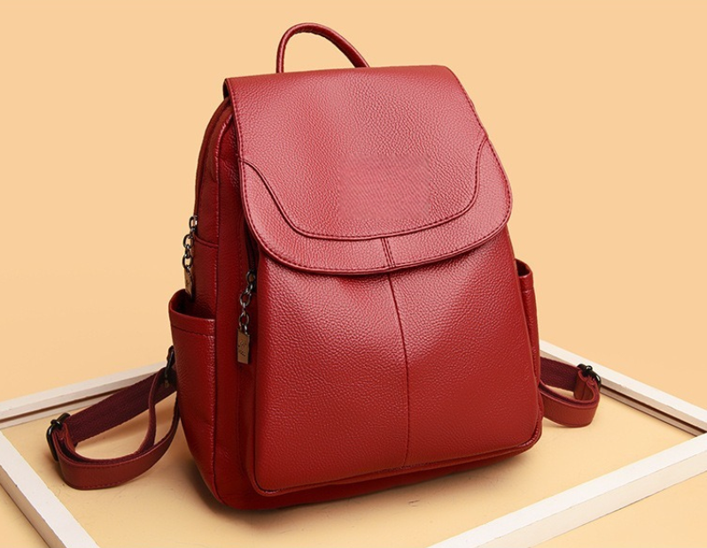 Sac à dos voyage rouge profond en effet cuir, silhouette élégante avec rabat avant, poches latérales et finitions soignées dans un cadre blanc et fond crème brulée