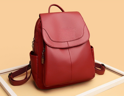 Sac à dos voyage rouge profond en effet cuir, silhouette élégante avec rabat avant, poches latérales et finitions soignées dans un cadre blanc et fond crème brulée