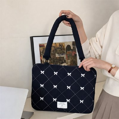 Sacoche ordinateur femme noir matelassée avec motifs nœuds blancs, tenue à la main sur un bureau, protection élégante pour PC portable.