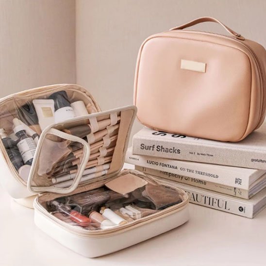 Trousse de toilette femme beige crème et rose poudré posée sur des livres, compartiments ouverts avec maquillage et soins, design élégant et féminin.